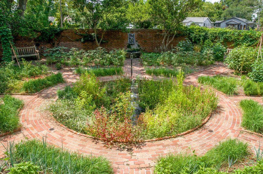 The Walled Garden at Longue Vue in New Orleans has a circular bed and brick pathways. Plants include an array of edible and medicinal plants like rosemary, anise hyssop, tulsi, celosia, okra, peppers, and hibiscus.