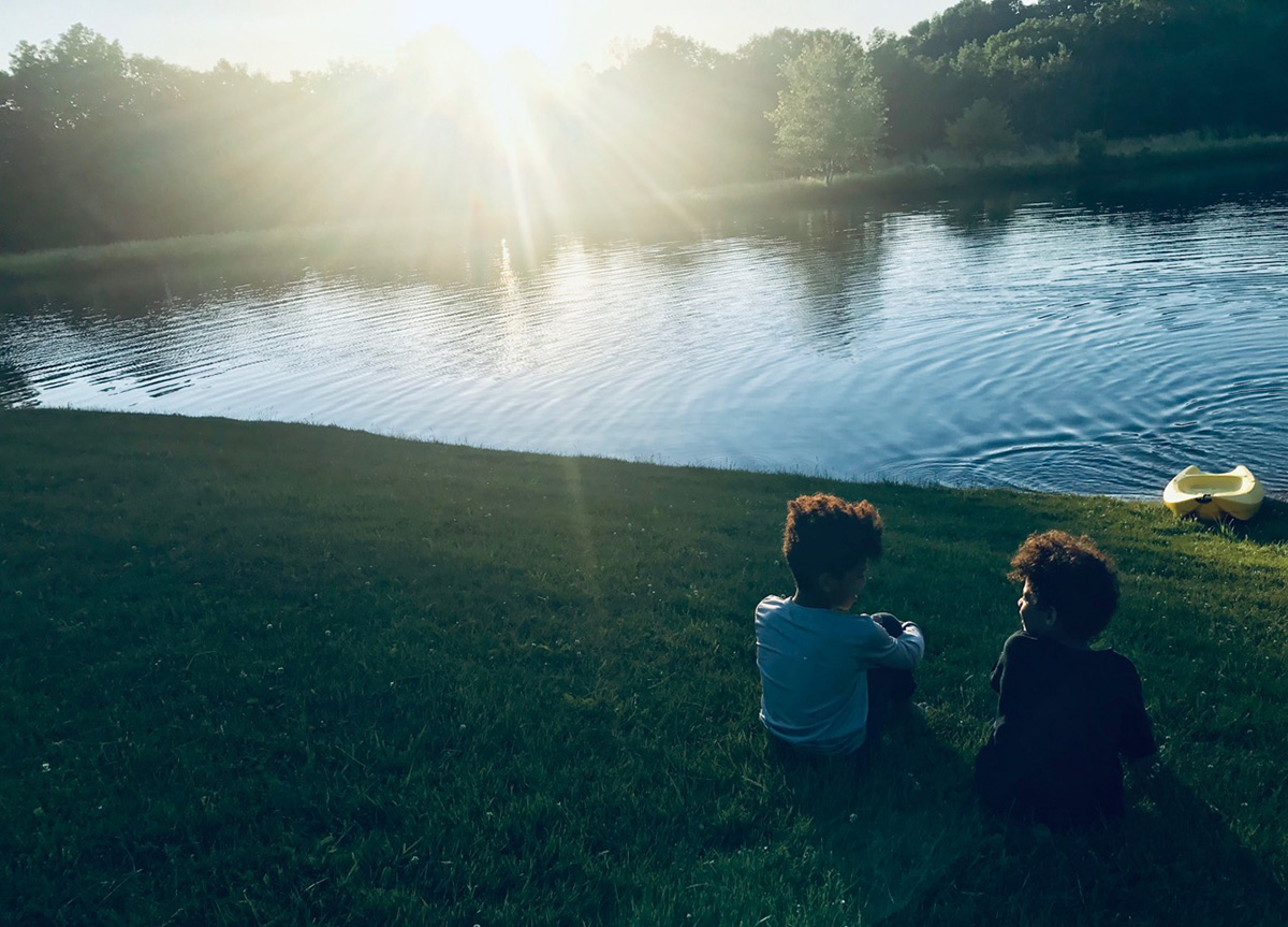 Two boys sit in a chemical-free lawn near a pond in the evening light.